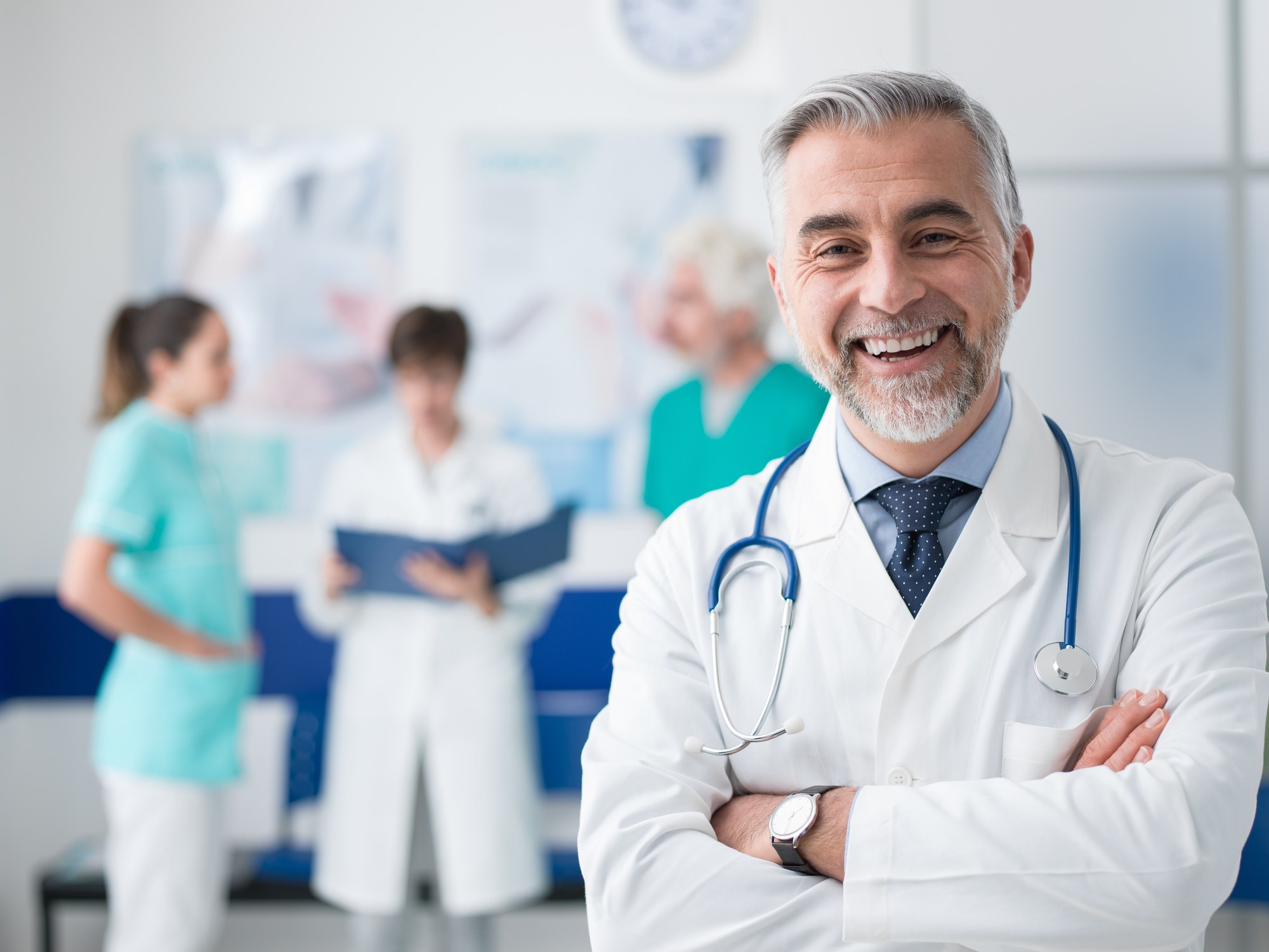 A smiling physician with arms crossed in a busy practice, staff and a patient in the background.